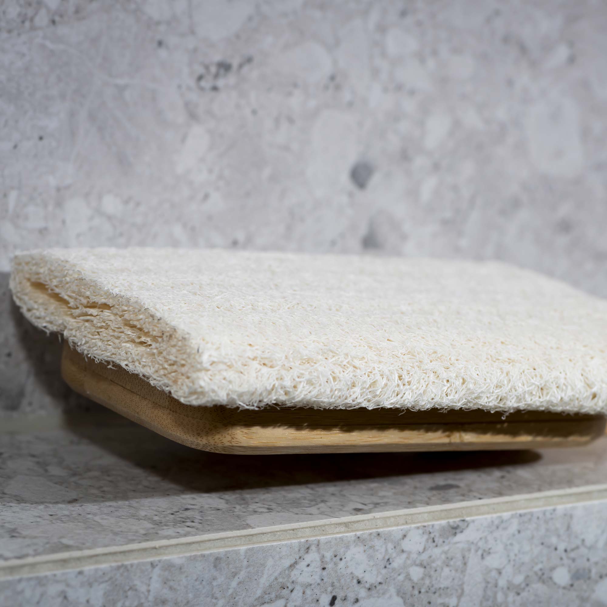 White bath mat with wooden border on a gray marble surface. Victoria James Body Loofah Brush - Square