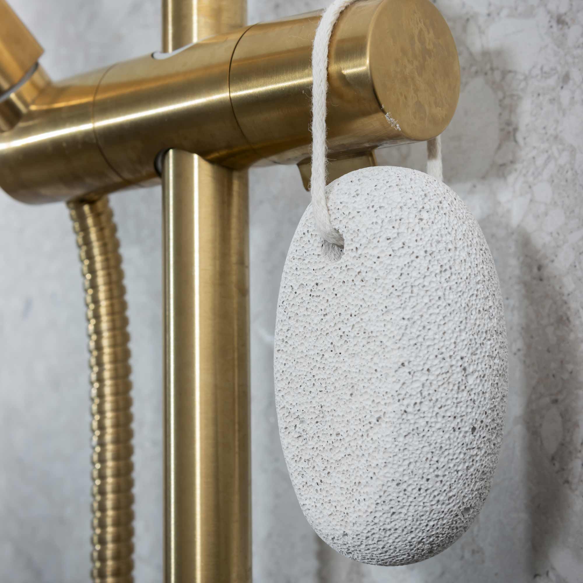 White pumice stone hanging from a gold shower head on a marble wall. Victoria James Body Pumice Stone