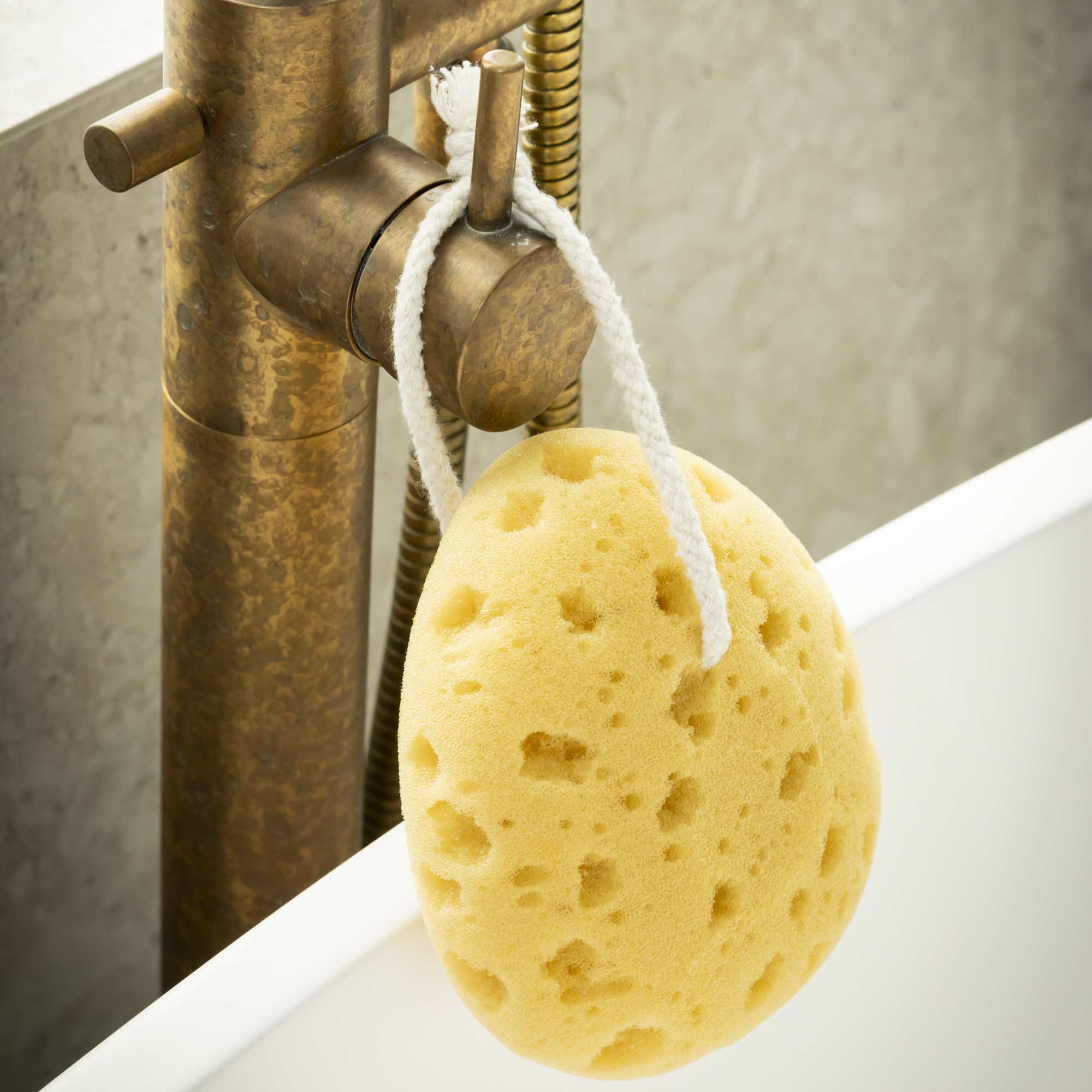 Yellow sponge hanging from a faucet in a bathroom setting. Victoria James Bath Sponge