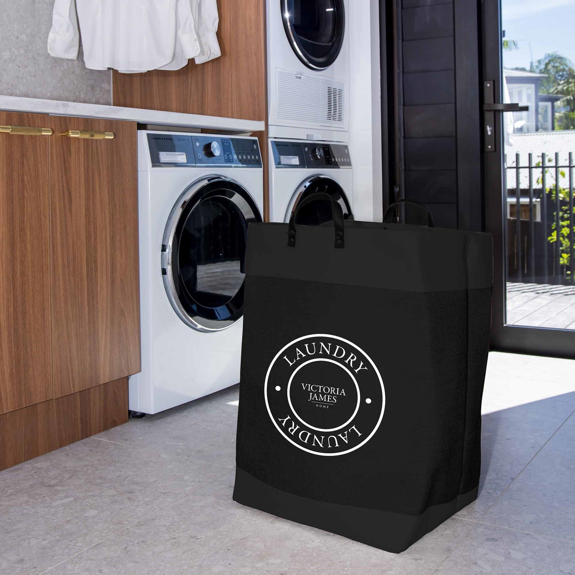 Black laundry bag with 'Victoria James' branding in a laundry room. Victoria James Home Laundry Hamper