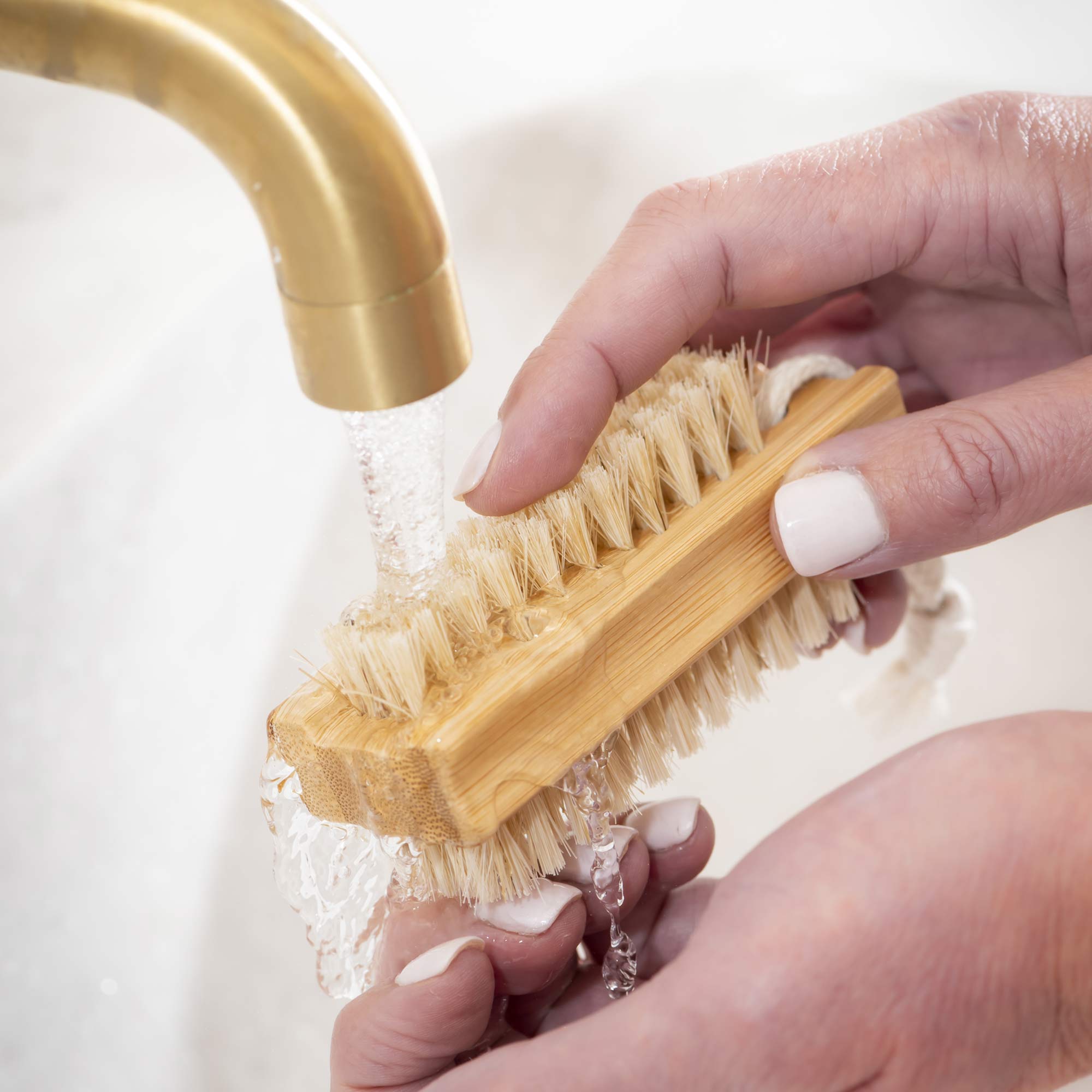 Hand holding a wooden brush under running water from a gold faucet. Victoria James Body Nail Brush