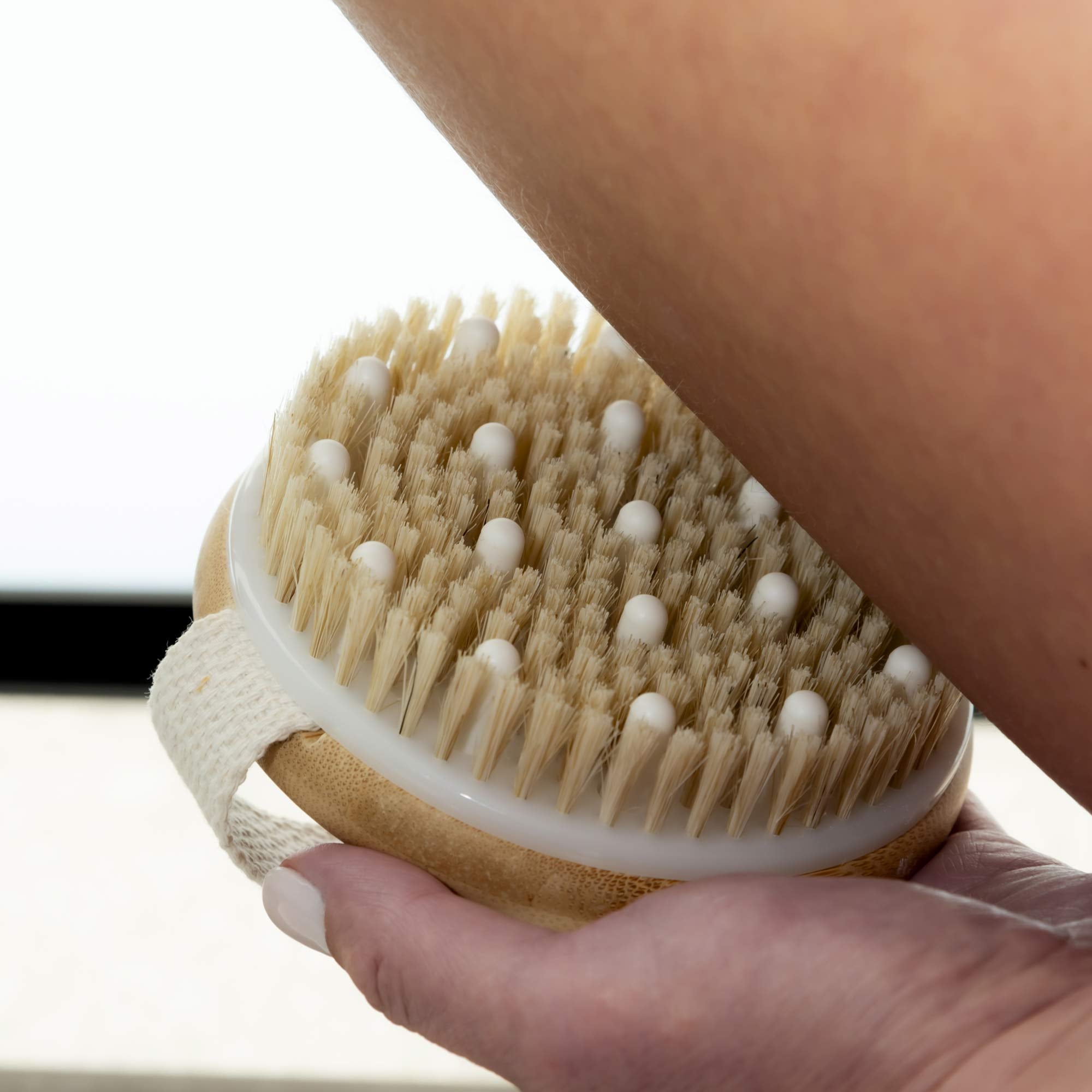 Person using a dry brush on their skin with a neutral background. Victoria James Massaging Body Brush