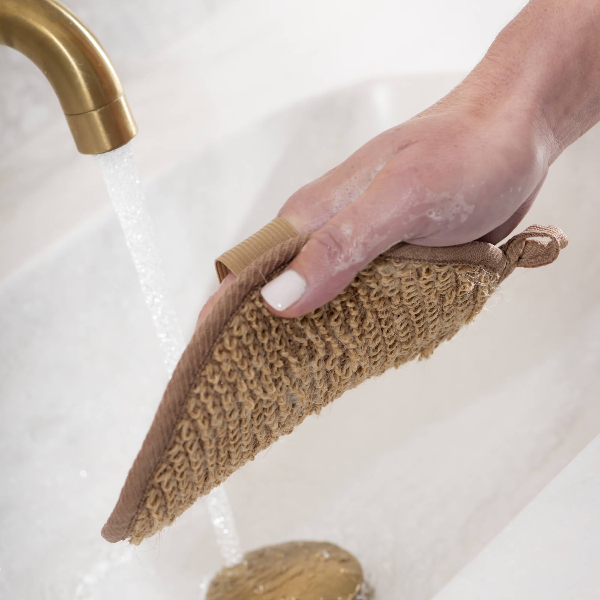 Hand holding a brown scrubber over a sink with water running. Victoria James Body Exfoliating Pad