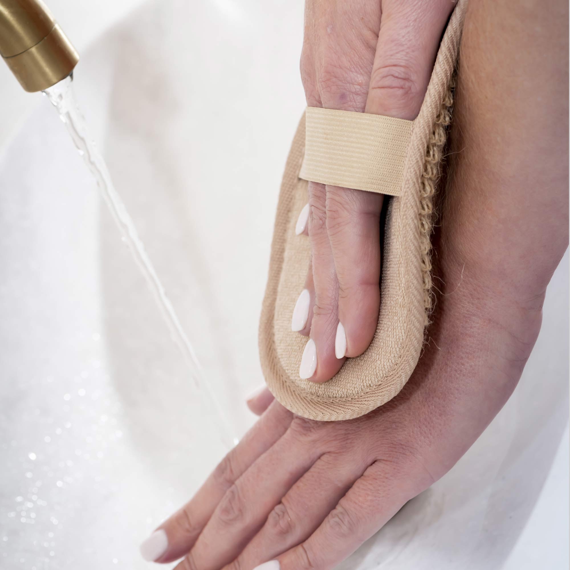 Hand holding a loofah with water flowing over it against a white background. Victoria James Body Exfoliating Pad